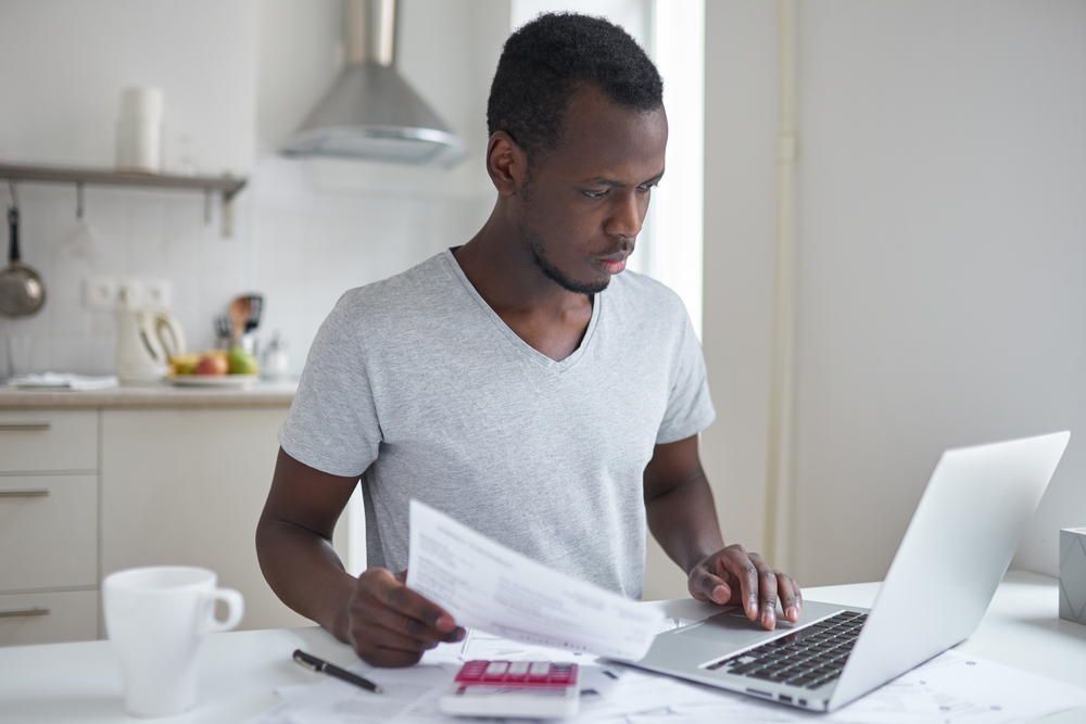 Jovem de frente para um laptop, segurando contas, preocupado com o pagamento da mensalidade da faculdade